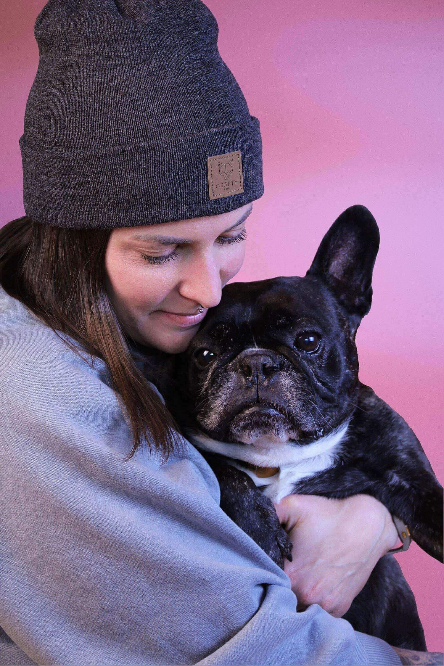 kerstin dörflinger mit ihrem hund einer schwarzen französischem bulldogge. sie hält ihren hund im arm vor rosa hintergrund.